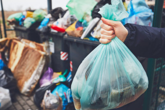 Photo of someone holding a full bag of rubbish in front of overflowing outdoor bins